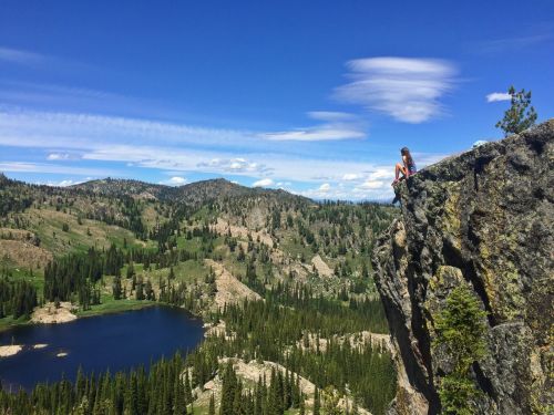 A person sits on a rocky ledge overlooking a lush valley and lake, surrounded by mountains and blue skies.