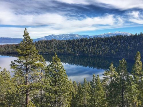 A serene lake surrounded by lush green trees and mountains under a cloudy sky. Reflections shimmer on the water's surface.