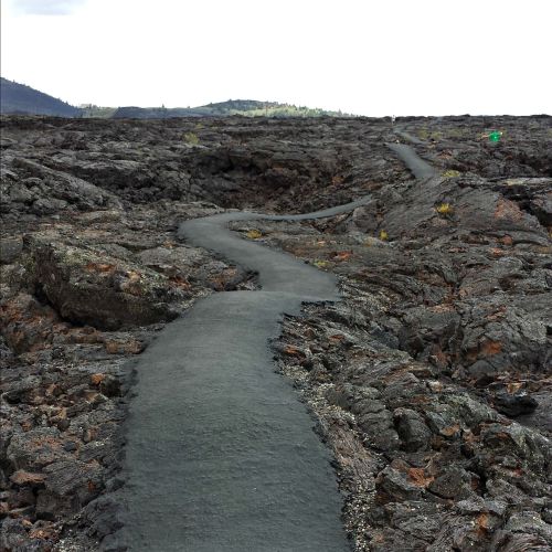 A winding path through a rugged lava field, surrounded by sparse vegetation and distant hills under a cloudy sky.
