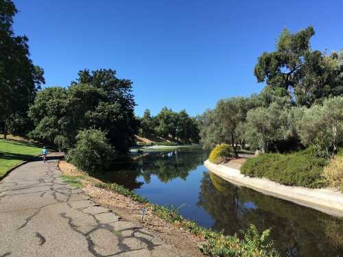 A serene park scene featuring a calm lake, lush greenery, and a walking path under a clear blue sky.