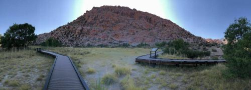 A panoramic view of a rocky mountain with a wooden pathway winding through a grassy area in the foreground.