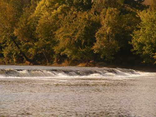 A serene river scene with a gentle waterfall and lush green trees lining the shore.