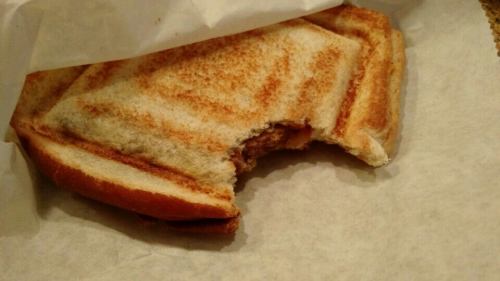 A partially eaten grilled cheese sandwich on white paper, showing golden-brown toasted bread and melted cheese.