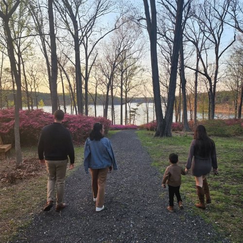 A family walks along a path by a lake, surrounded by trees and blooming flowers, during sunset.