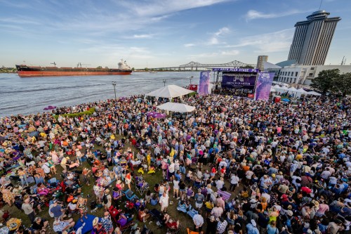 A large crowd gathers at a riverside festival, with a stage and a ship in the background under a clear blue sky.