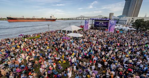 A large crowd gathers at a riverside festival with a stage, tents, and a cargo ship in the background.