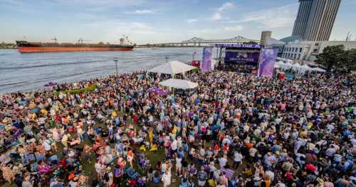 A large crowd gathers at a riverside festival with a stage, tents, and a cargo ship in the background.