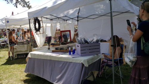A bustling outdoor market with white tents, vendors displaying goods, and visitors browsing and shopping.