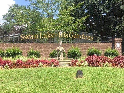 Sign for Swan Lake Iris Gardens with a fountain and colorful plants in front, set against a sunny backdrop.