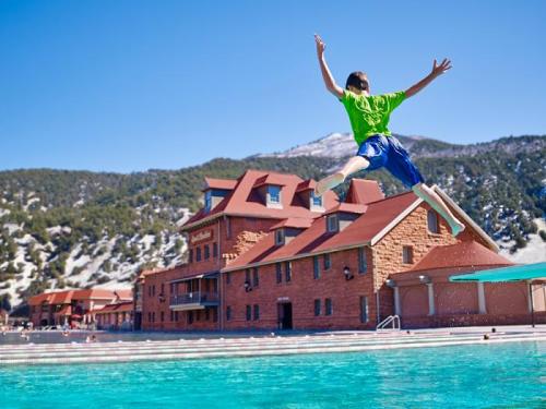 A child in a green shirt jumps joyfully into a pool, with a historic building and mountains in the background.
