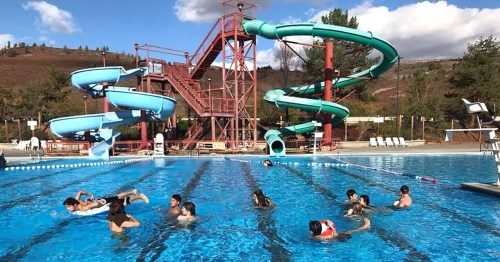 A sunny outdoor pool with water slides, featuring children swimming and playing in the water.