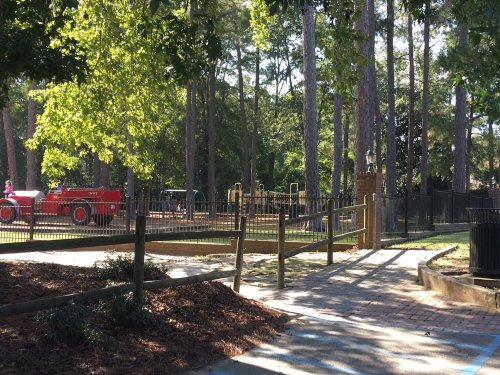 A park scene with a playground in the background, surrounded by tall trees and a fenced area.