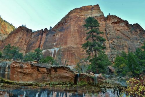 A towering red rock formation surrounded by trees and a small waterfall in a scenic landscape.