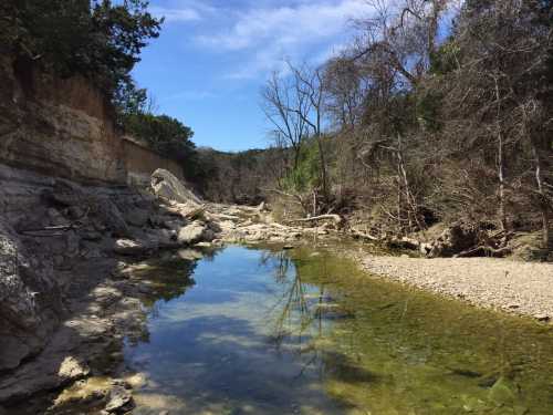 A serene riverbank scene with clear water reflecting trees and blue sky, surrounded by rocky terrain and dry vegetation.