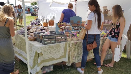 A market stall filled with colorful bath products, with people browsing and shopping in a sunny outdoor setting.