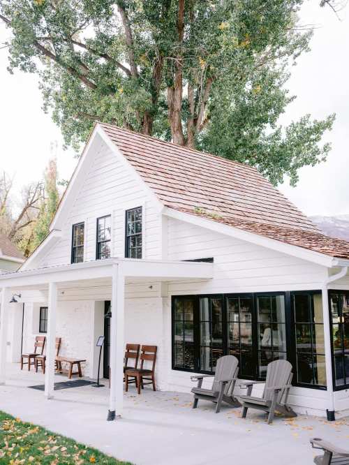 A charming white house with a sloped roof, large windows, and a tree growing nearby, featuring outdoor seating.