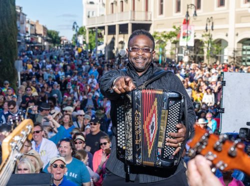 A smiling musician plays the accordion on stage, pointing to the crowd at a lively outdoor festival.