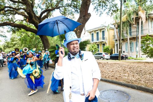 A man in a white suit and blue hat holds an umbrella while leading a colorful parade down a tree-lined street.