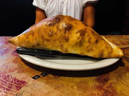 A large, golden-brown calzone sits on a plate with a knife, with a person in a white shirt in the background.