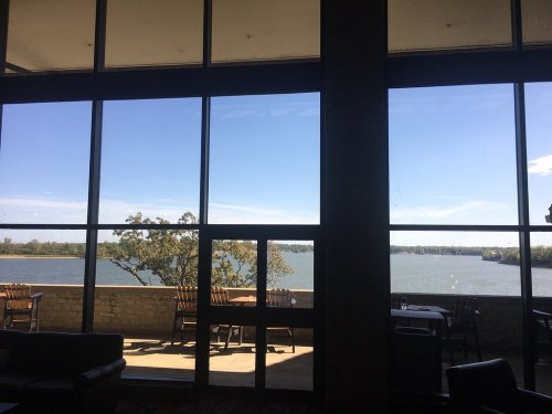 View of a serene lake through large windows, with trees and blue sky visible in the background.