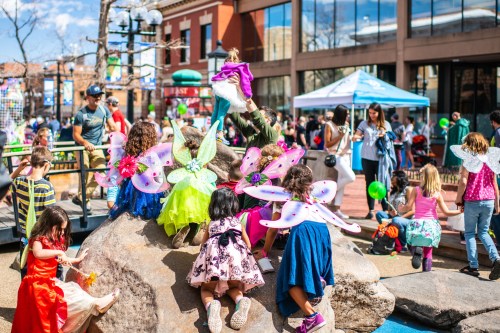 Children in colorful fairy costumes play on a rock in a lively outdoor setting, with families and activities in the background.