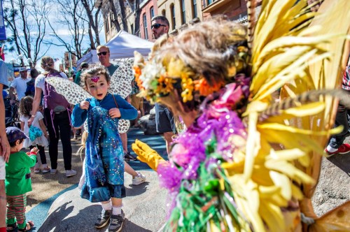 A child in a blue dress with wings interacts with a colorful performer at a lively outdoor festival.