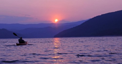A person kayaking on a calm lake at sunset, with mountains silhouetted in the background and a purple sky.