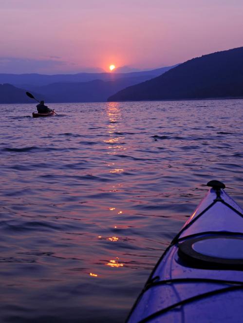 A serene sunset over a lake, with a kayaker paddling in the foreground and mountains in the background.