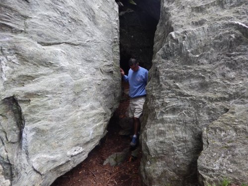 A person navigating through a narrow gap between two large rock formations in a forested area.