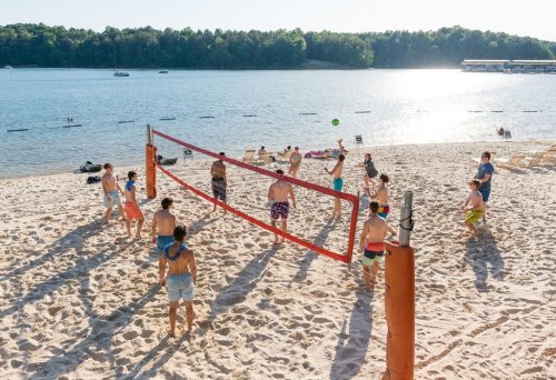 A group of people playing beach volleyball on a sandy shore by a lake, with boats in the background and trees lining the shore.