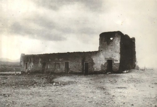 Black and white photo of a weathered, abandoned stone building with a partially collapsed wall and cloudy sky.