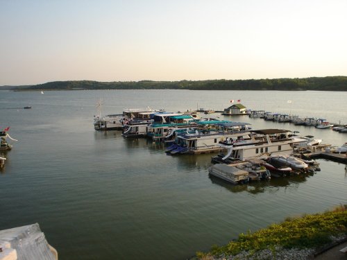 A serene marina with houseboats and small boats on calm water, surrounded by green hills under a clear sky.