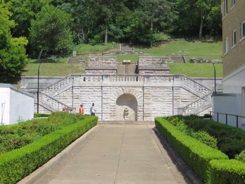 A stone staircase leads up a hill, flanked by greenery, with two people walking towards an archway at the top.
