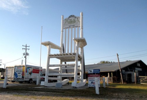 A large white rocking chair stands in front of a building, with signs and a U-Haul truck nearby.