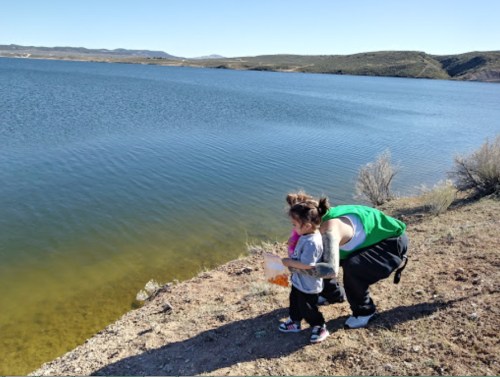 A child and an adult stand by a lake, looking at the water with hills in the background under a clear blue sky.