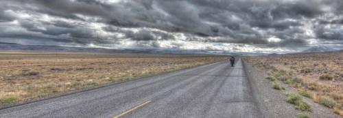 A lone motorcyclist rides down a wide, empty road under a dramatic sky filled with clouds.