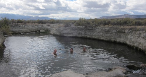Three people enjoy a natural hot spring surrounded by rocky terrain and sparse vegetation under a cloudy sky.