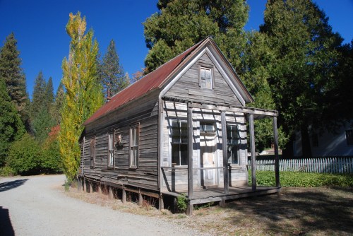 A weathered wooden house with a red roof, surrounded by trees and a gravel path.