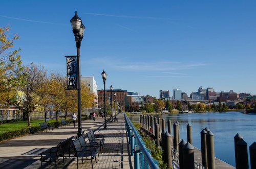 A scenic waterfront walkway lined with benches and lampposts, with city buildings in the background under a clear blue sky.