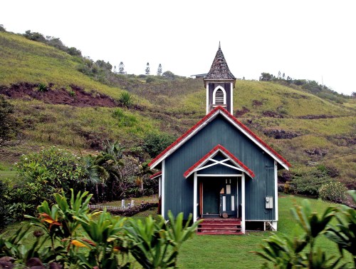 A small, green church with a red roof sits on a grassy hillside, surrounded by lush vegetation.