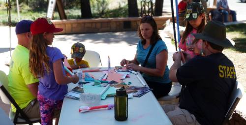 A group of children and adults sit at a table, engaged in a craft activity outdoors.