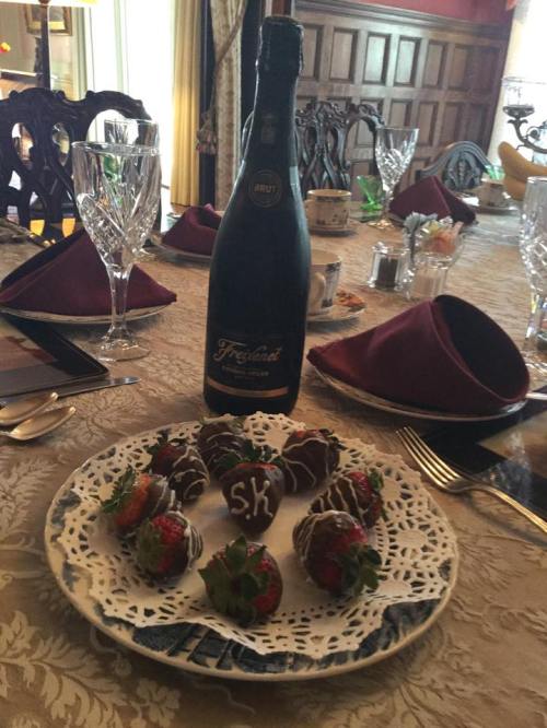 A plate of chocolate-covered strawberries with initials, accompanied by a bottle of sparkling wine on a beautifully set table.