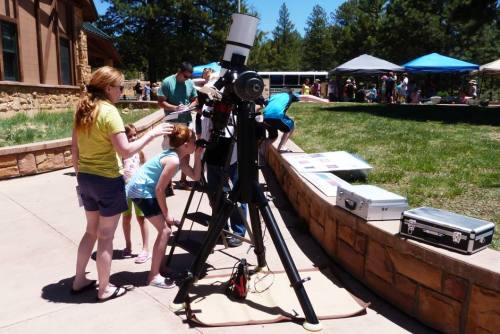 A group of people, including children, observe a large telescope set up outdoors at a sunny event.