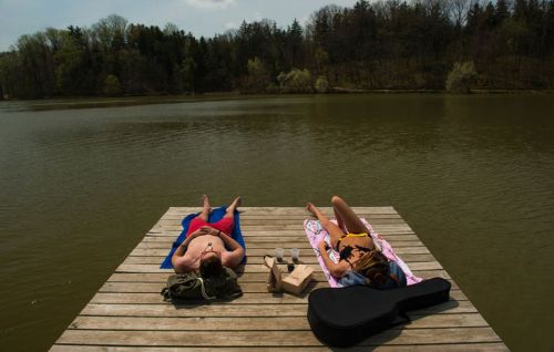 Two people sunbathe on a dock by a calm lake, surrounded by trees, with a guitar and drinks nearby.