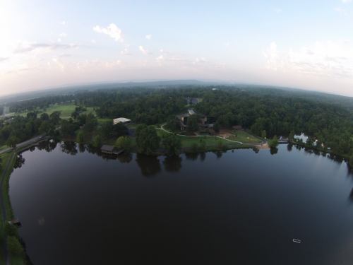 Aerial view of a serene lake surrounded by lush greenery and distant hills under a cloudy sky.