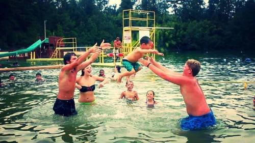 A group of children and teens play in a lake, with one boy jumping into the water while others cheer and splash around.
