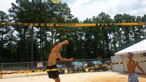 Two young men playing beach volleyball, one jumping to hit the ball over the net, surrounded by trees and a sandy court.