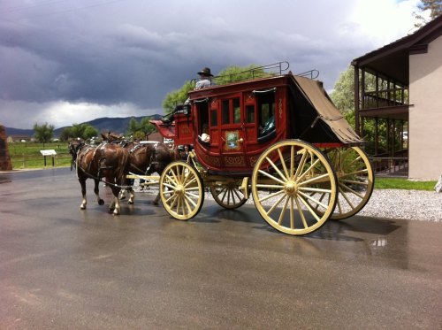 A vintage horse-drawn stagecoach parked on a wet road, with a driver seated on top and dark clouds in the background.