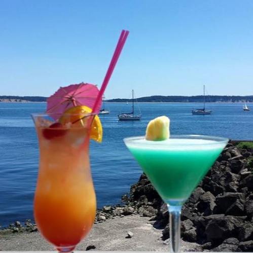 Two colorful cocktails with umbrellas sit on a table by the water, with sailboats in the background and a clear blue sky.