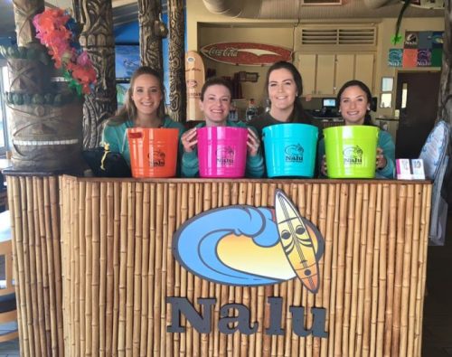 Four women stand behind a bamboo bar at Nalu, each holding colorful buckets, smiling at the camera.